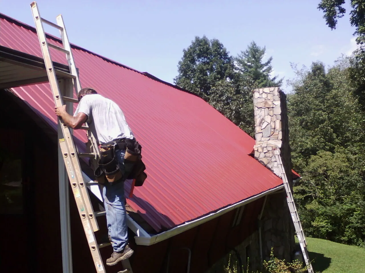Roofer on a ladder beside a red metal roof and stone chimney — Soffit & Fascia Repair by Roofing Brothers in Paducah